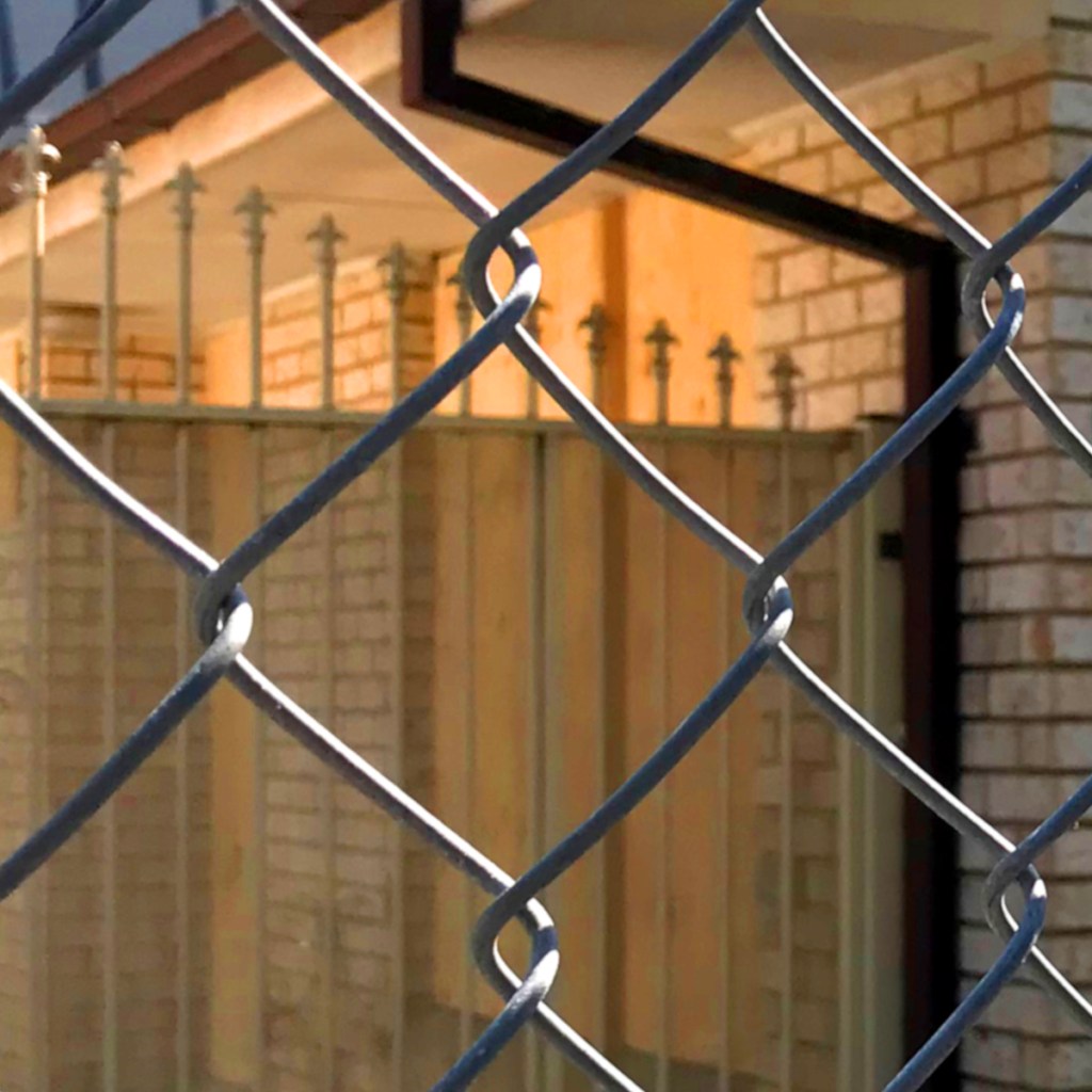 Close-up of a chain-link fence with blurred wooden structures and brick walls visible in the background.