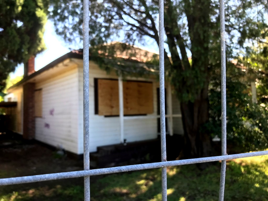 A boarded-up house seen through a metal fence, surrounded by trees and grassy area.