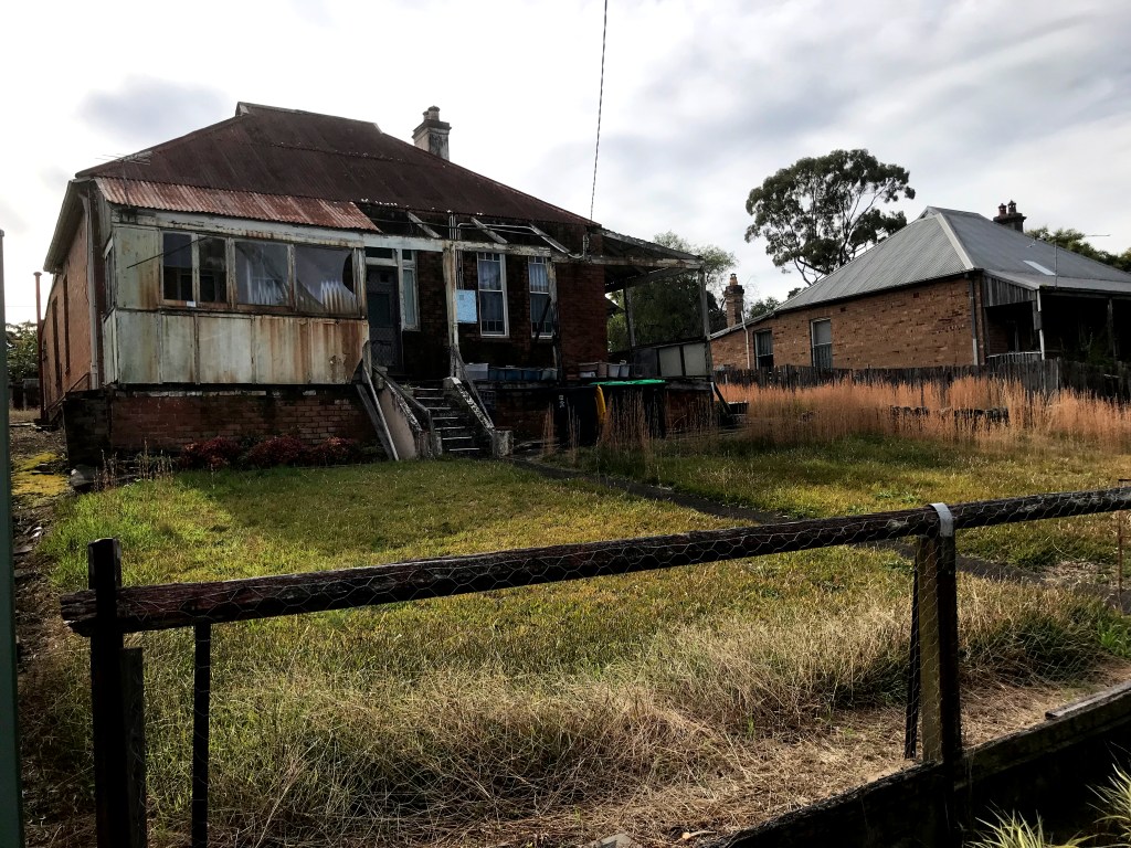 A derelict house with a weathered facade, overgrown grass, and a visible fence, indicating abandonment.