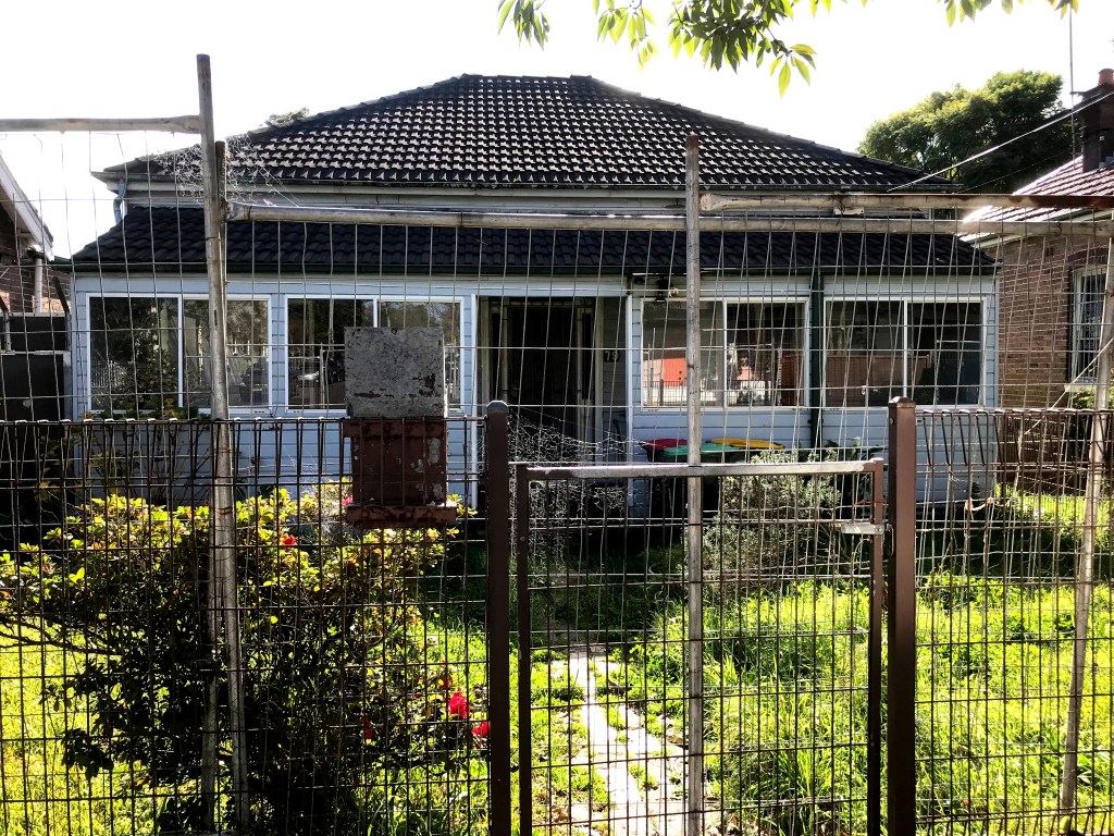A single-storey house surrounded by a wire fence, with a garden featuring green grass and flower beds in front.