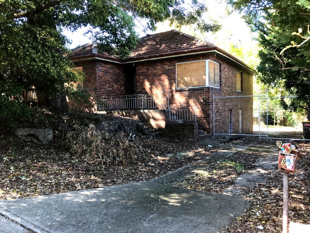 Abandoned brick house with boarded windows, surrounded by overgrown vegetation and a fence.