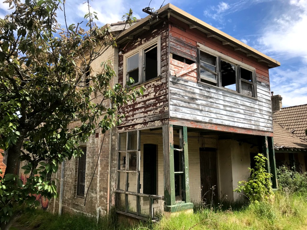 Exterior view of an abandoned house with peeling paint and boarded-up windows, surrounded by tall grass and trees.