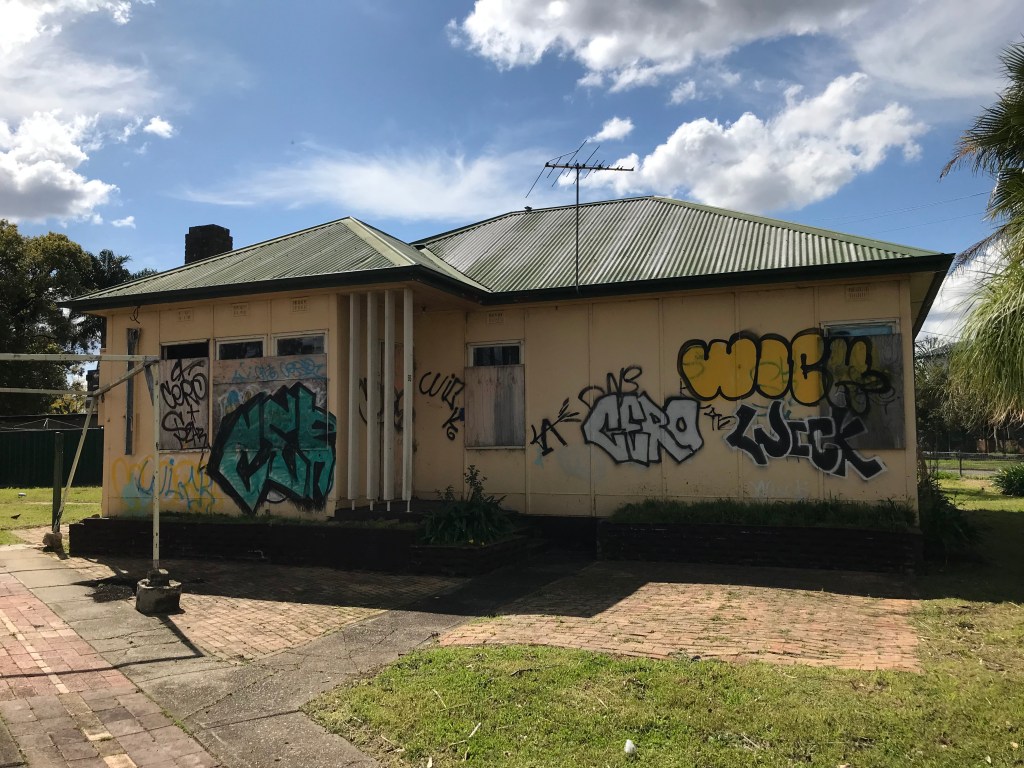 A vacant, graffiti-covered house with boarded windows and a green roof, situated in a grassy area under a blue sky.