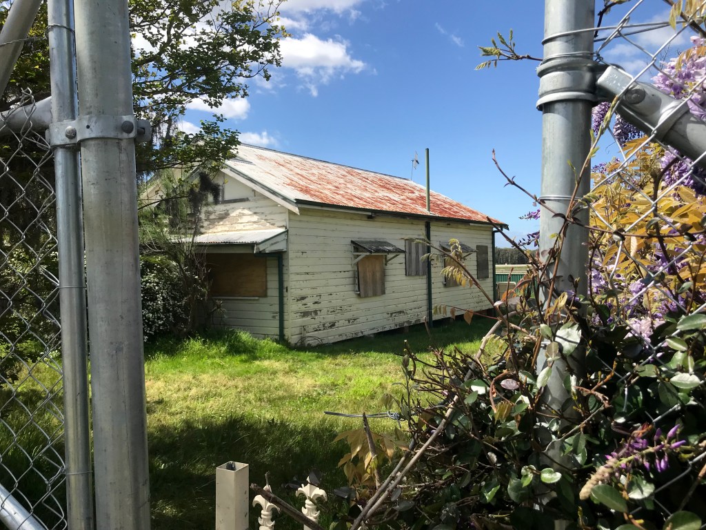 An abandoned house with boarded windows and overgrown grass, viewed through a metal fence.