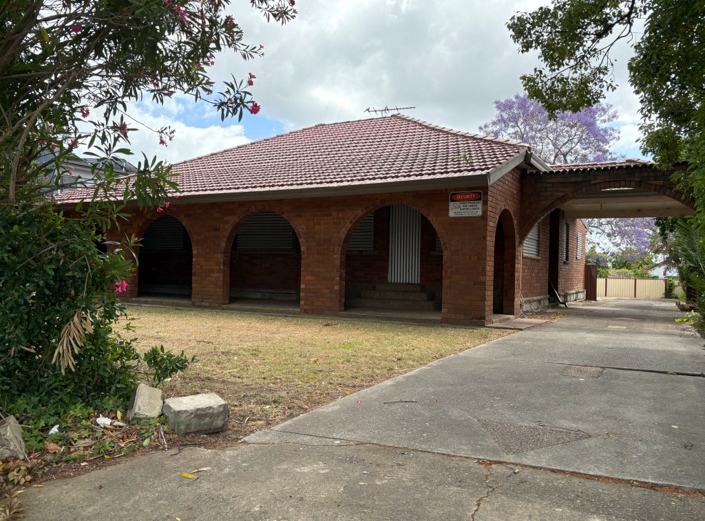 A brick house with a red tiled roof, featuring arched doorways and shuttered windows, set in a grassy area with some shrubs and nearby trees.