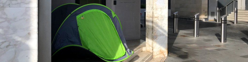 A green and blue tent positioned at the entrance of a modern building, with a marble wall on one side and metallic bollards in the background.