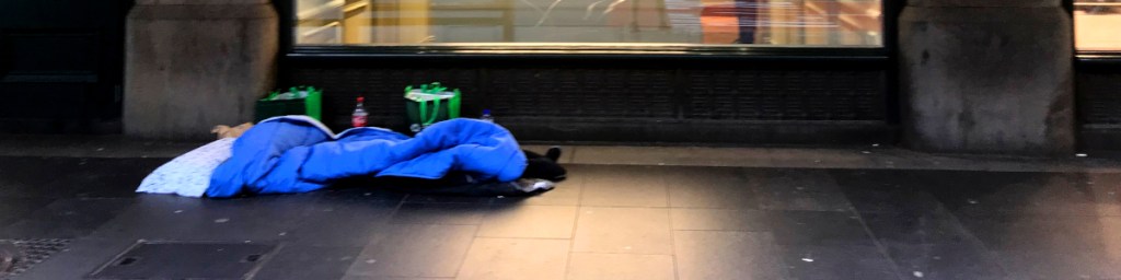 A blue sleeping bag laid out on the pavement in front of a shop window, with bags placed nearby.