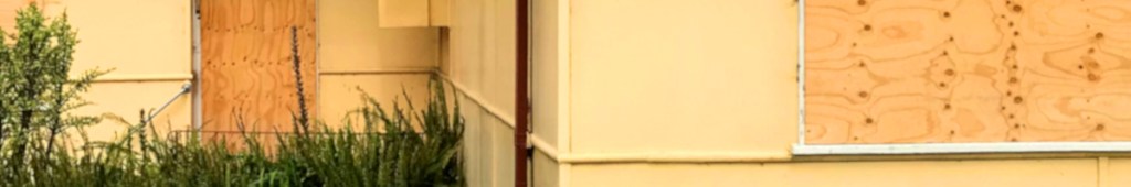 Exterior of a building featuring a boarded-up door and window, with yellow walls and surrounding greenery.