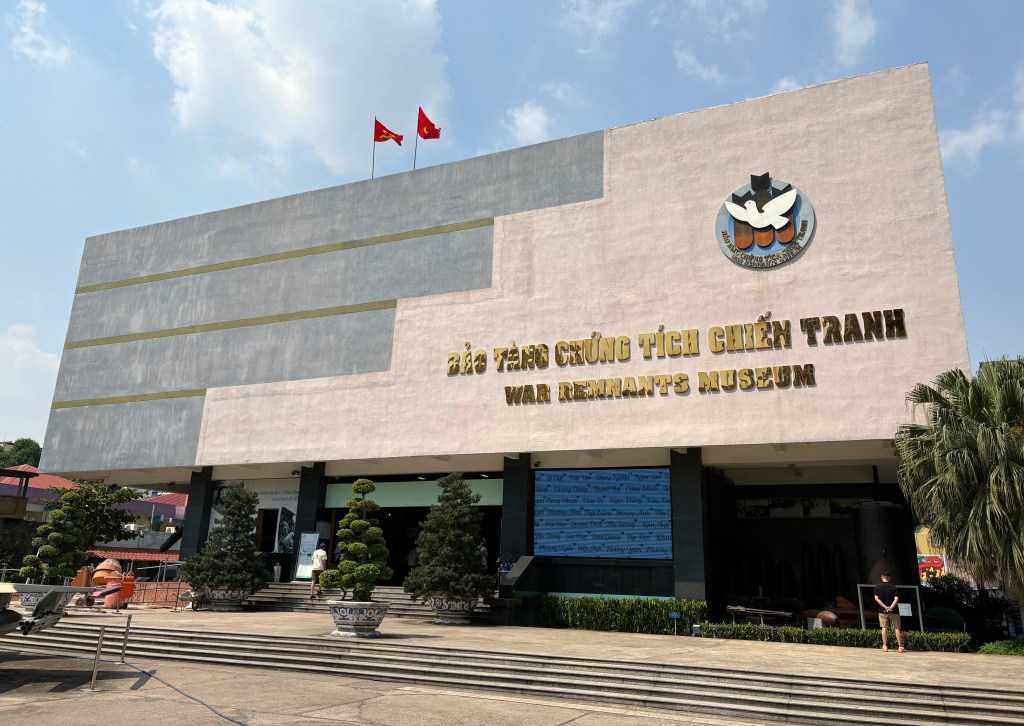 Exterior view of the War Remnants Museum in Ho Chi Minh City, featuring a modern building design with clear blue skies and greenery in the foreground.