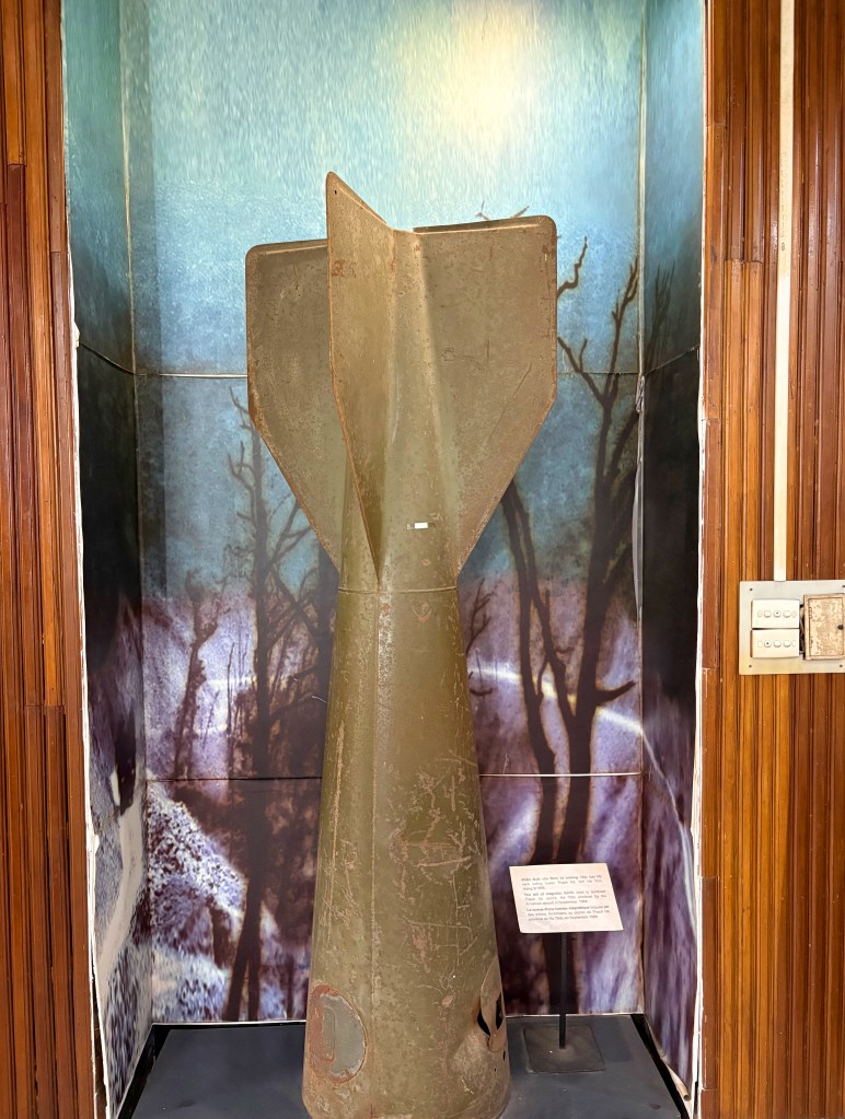 A large, rusted bomb shell displayed in a museum, set against a backdrop of dark trees and a misty landscape.