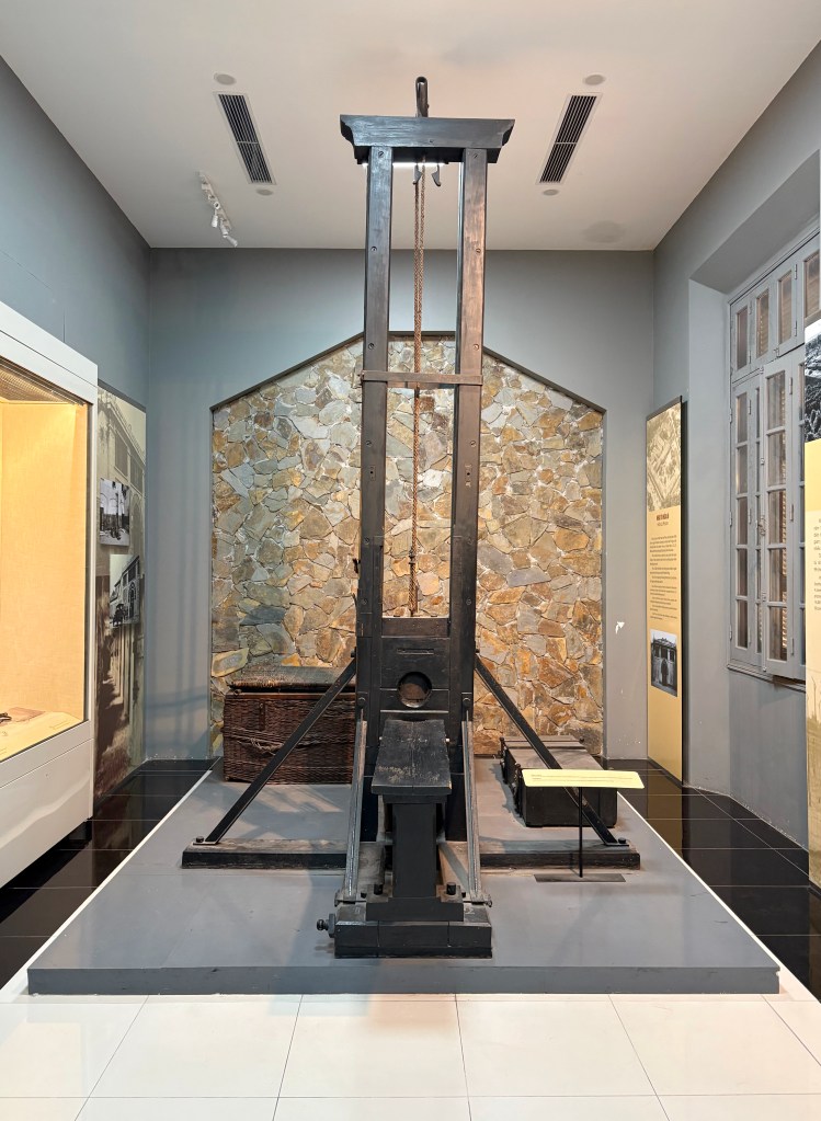 A historical guillotine displayed in a museum setting, with a stone wall backdrop and exhibit information visible nearby.