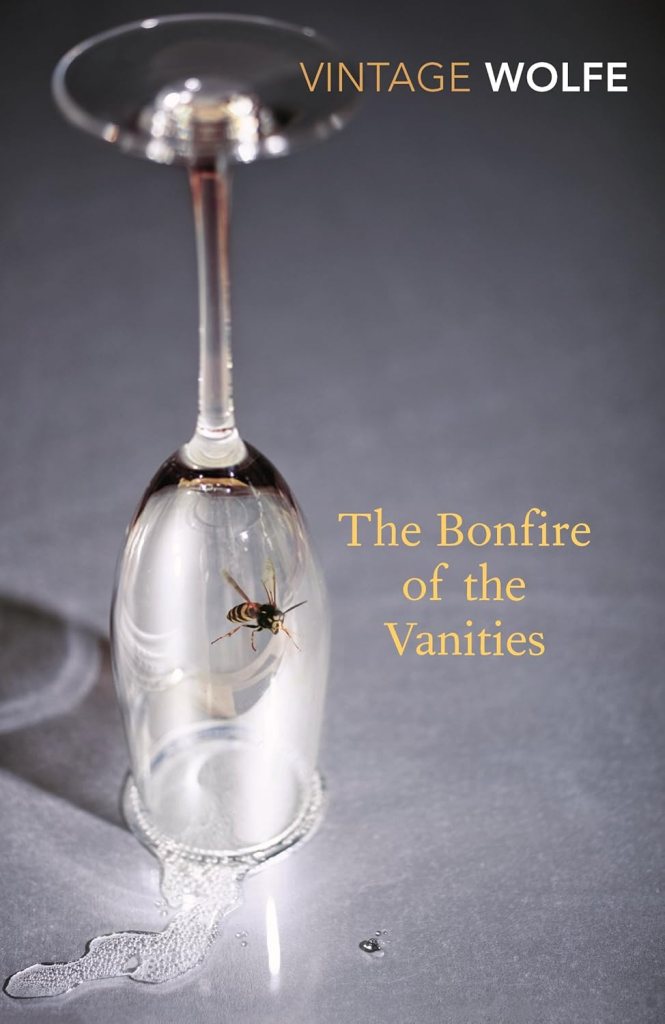 A close-up of a wine glass turned upside down with a bee trapped inside, resting on a reflective surface with droplets of liquid around it. The title 'The Bonfire of the Vanities' and the text 'VINTAGE WOLFE' are visible above the glass.