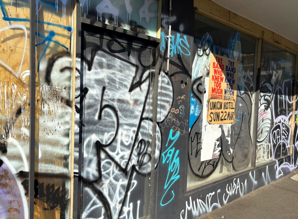 A street view of a wall covered in graffiti with a poster for a band event at the Union Hotel on 22 March. The poster includes bold text and is surrounded by various colourful graffiti and scribbles.