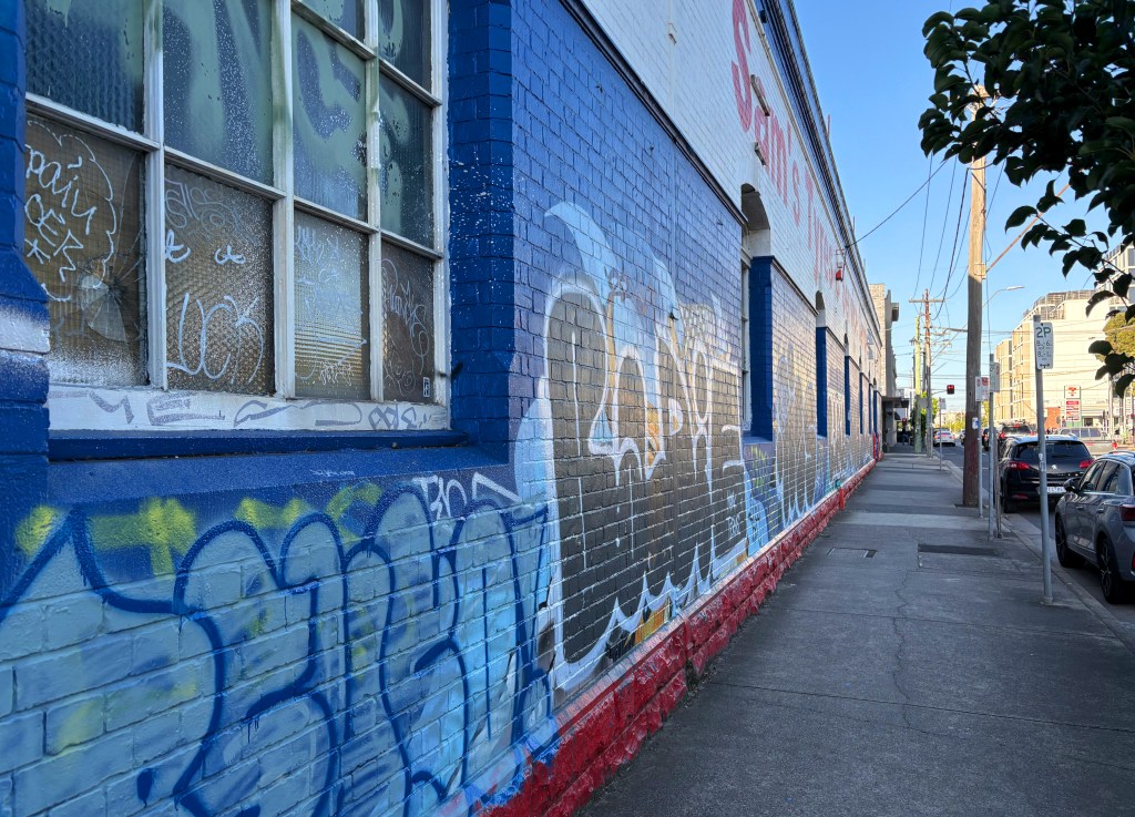 A vibrant blue wall decorated with various graffiti artworks, featuring a mix of colorful tags and designs, adjacent to a sidewalk with parked cars and a clear sky.