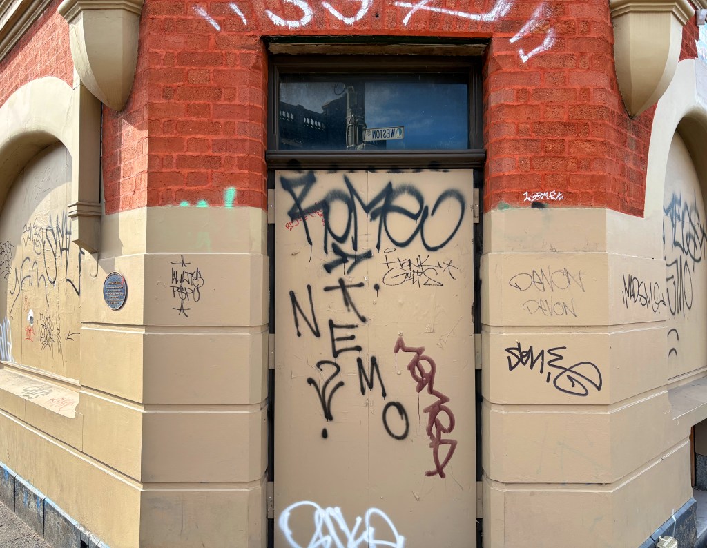A doorway covered in various graffiti, located on a red-bricked building. The surface features multiple tags and scribbles in different styles and colours.