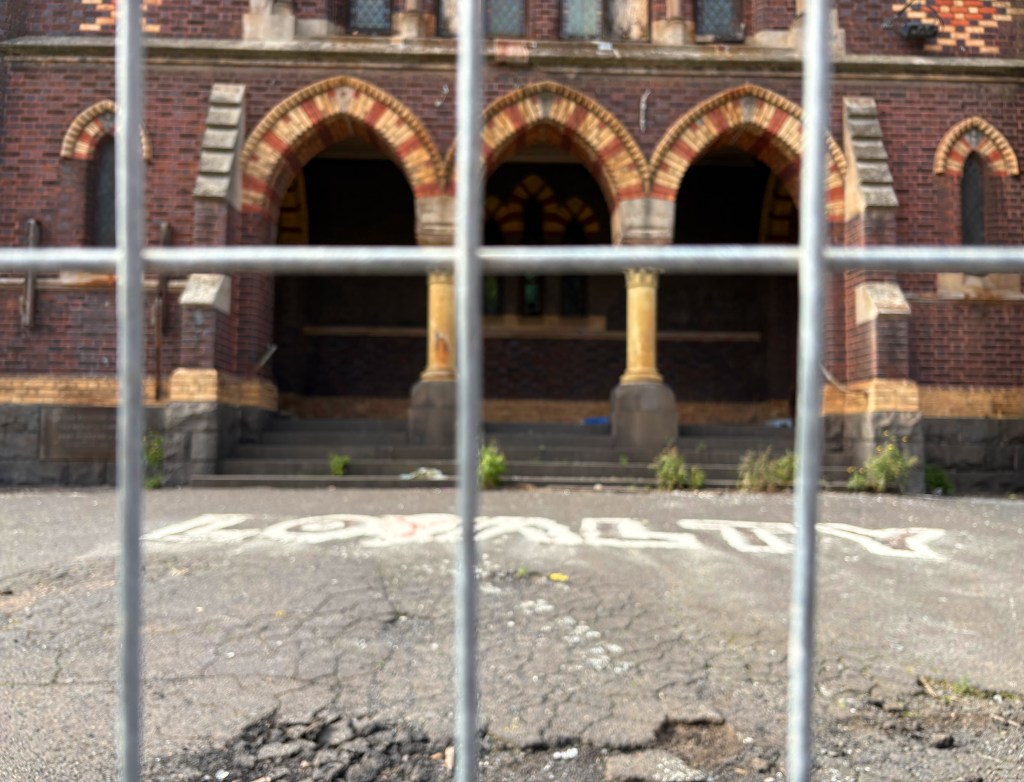 View of an old brick building with arched windows, partially obscured by a fence. The pavement in front shows a faded word 'LOYALTY' painted on it, with weeds and cracks visible.