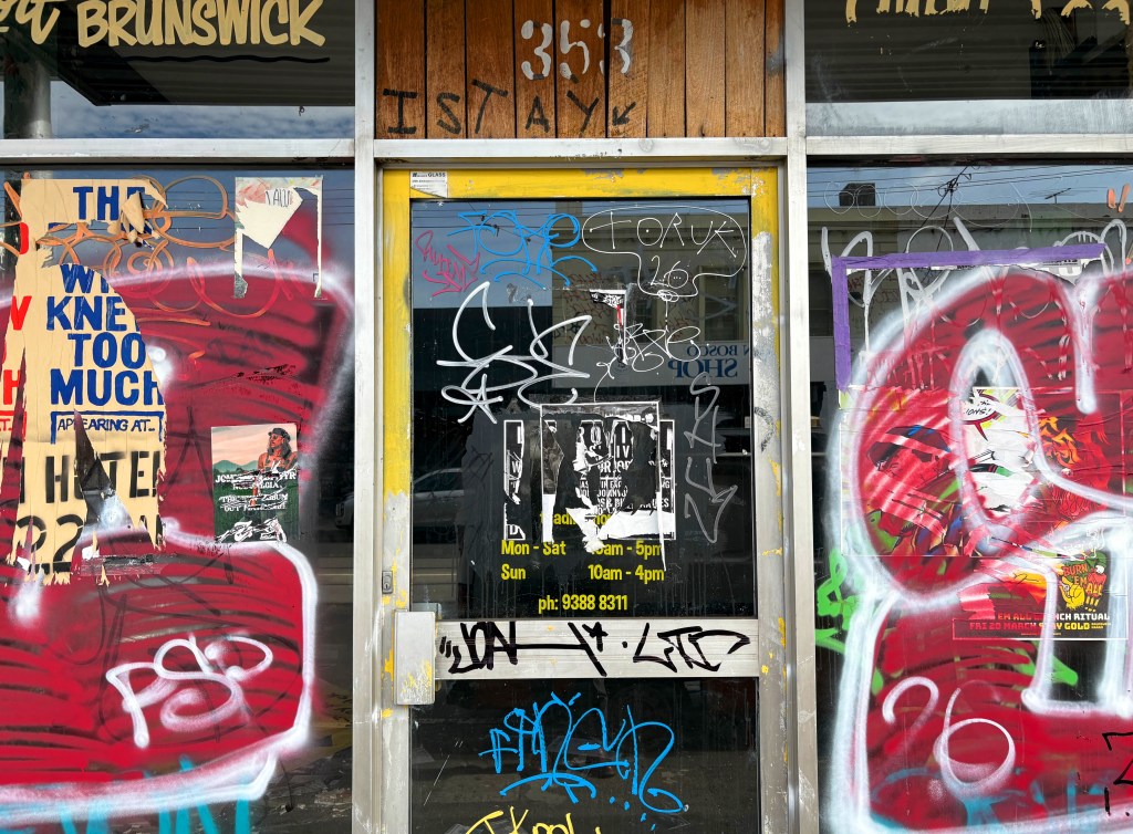 A close-up of a shopfront in Brunswick covered in graffiti, featuring various posters and a partially obscured sign indicating business hours.