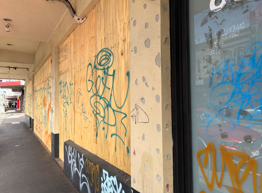 A street view showing a boarded-up building with wooden planks and colourful graffiti on the walls, with a glimpse of shops in the background.