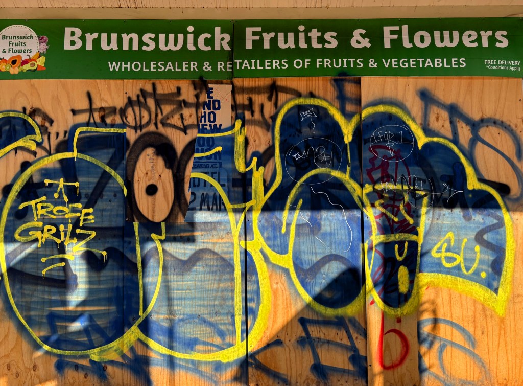 A close-up of the Brunswick Fruits & Flowers sign covered in graffiti on a wooden board, with yellow and blue spray paint obscuring the original text.