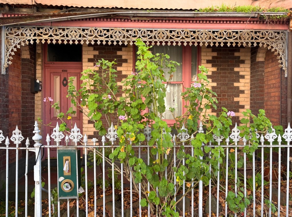 A vintage-style house front with intricate ironwork detailing, featuring a reddish-brown brick facade and a pink door numbered 601. A flowering plant grows near the entrance, alongside a white picket fence with a green letterbox.