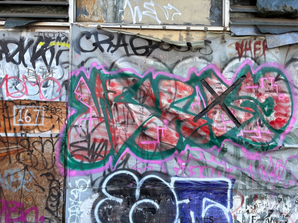 A wall covered in vibrant graffiti, featuring overlapping tags and colourful designs, with the prominent tag 'JBS' in bright pink and green.