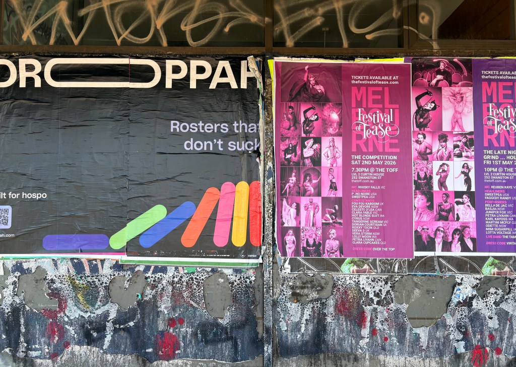 A street view of two posters on a wall. The left poster promotes a service with the tagline "Roster that don't suck" and includes a colourful design. The right poster advertises the 'Mel Festival of Tease' event, featuring details about a competition on 2nd May 2026, with vibrant pink backgrounds and images of performers. Graffiti and peeling paint are visible on the wall.