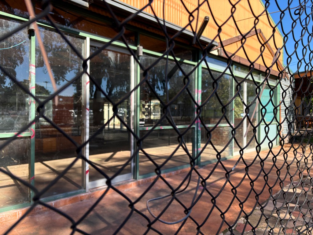 A view through a chain-link fence of the front of a building with large glass windows, reflecting trees and a clear blue sky.