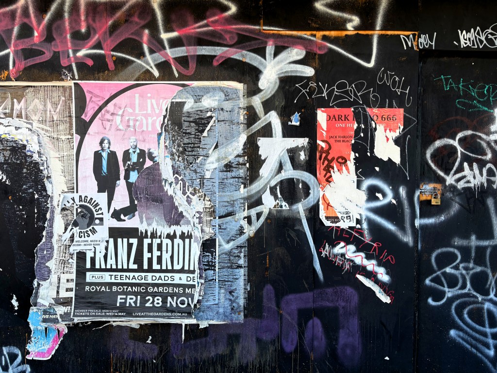 A wall covered in various layers of posters and graffiti, including an advertisement for a Franz Ferdinand concert, alongside other torn flyers and colourful graffiti tags.