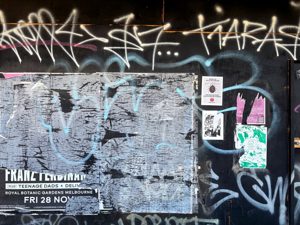 A wall covered in various graffiti and posters, featuring a large black area with missing sections, several colourful flyer posters, and overlapping spray paint graffiti.