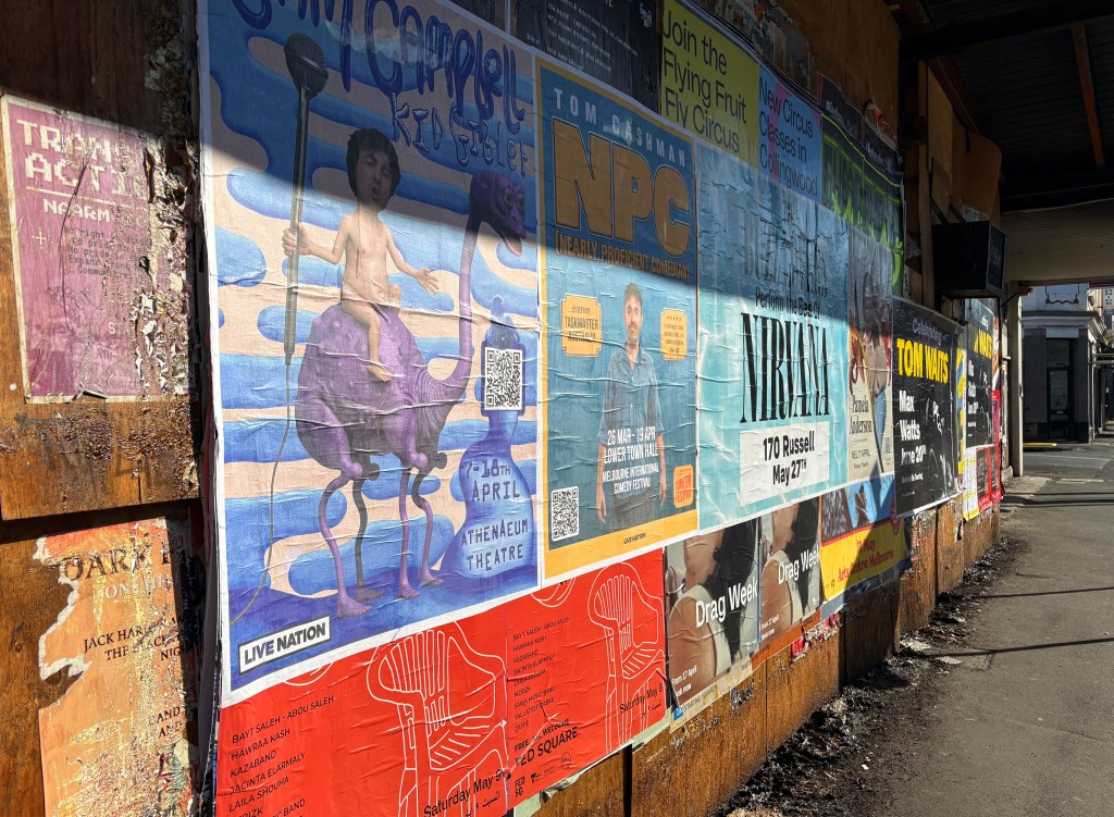 A wall covered with vibrant posters advertising various events and performances, including a comedy show and a circus. The posters feature bright colours and artistic designs, with some mentioning specific dates and venues.