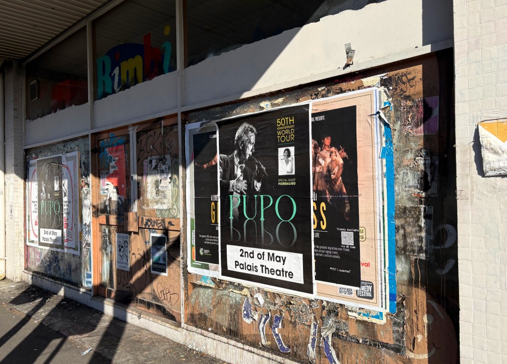 A weathered wall displaying various concert posters, including one for the artist Puopo, advertising a performance on 2nd May at the Palais Theatre. The area shows signs of wear and graffiti.