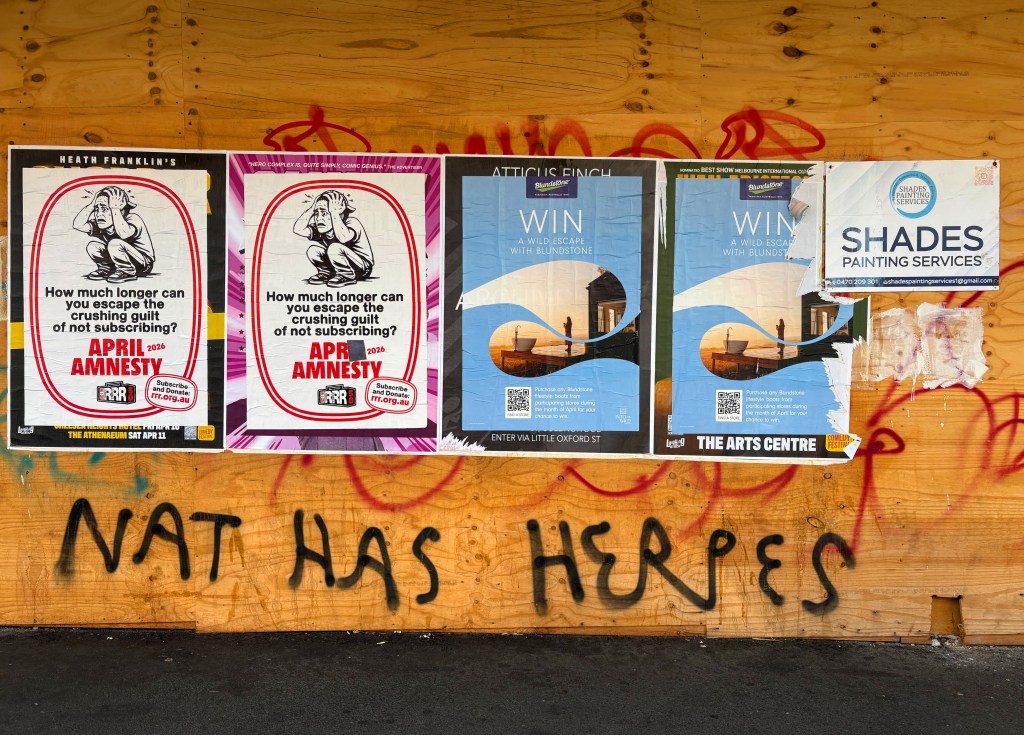 A wooden wall covered with various posters promoting events and services, including advertisements for 'Amnesty' and 'Win a wild escape with Blundstone', alongside graffiti reading 'NAT HAS HERPES'.