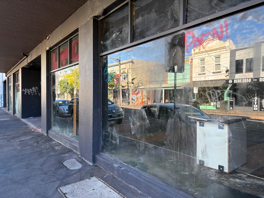 A partially obscured view of a street with shopfronts featuring dirty glass windows and graffiti, reflecting parked cars and buildings across the road.