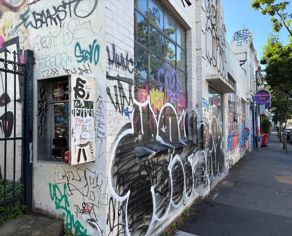 A street view of a building covered in vibrant graffiti, featuring various tags and designs on its walls. There is an entrance with a display cabinet and a sign for a nearby business visible.