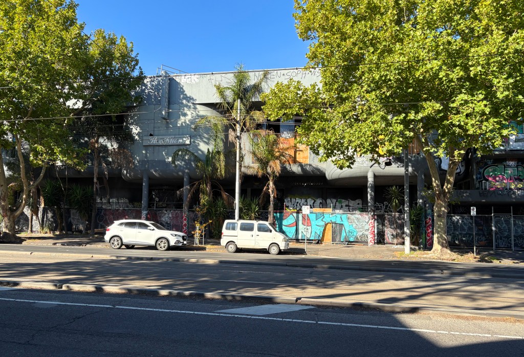 Abandoned building with graffiti on the exterior, surrounded by trees and parked cars on a sunny day.