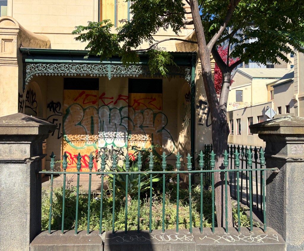 A rundown building with boarded-up windows covered in graffiti, surrounded by a green fence and overgrown vegetation, with trees partially in view.