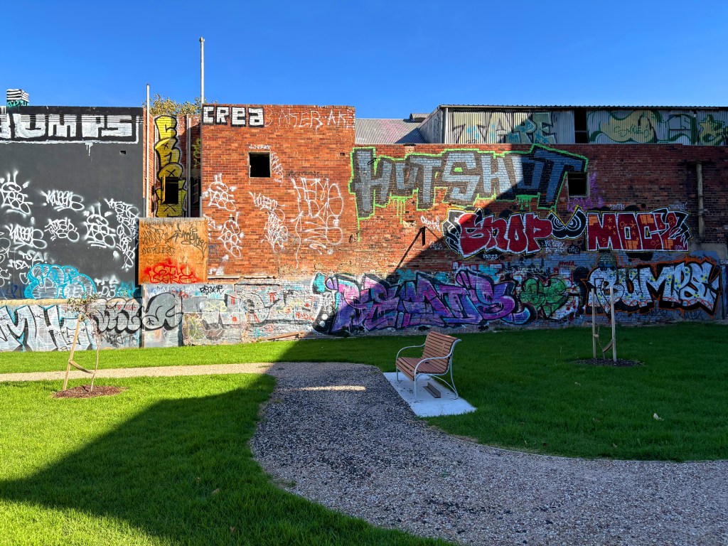 A mural-covered brick wall featuring vibrant graffiti art in various colours and styles, alongside a small park area with green grass and a gravel path.