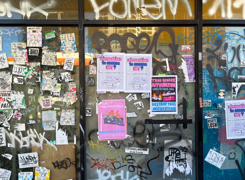 A glass window covered with various posters, stickers, and graffiti, featuring messages related to social issues and activism.