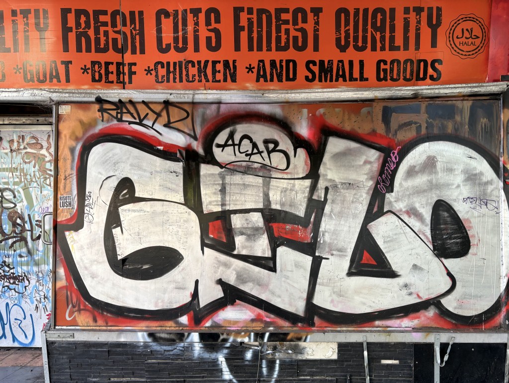 A colourful graffiti mural depicting the word 'GILO' on a metal board, set against a background of various other graffiti and advertising for meat products.