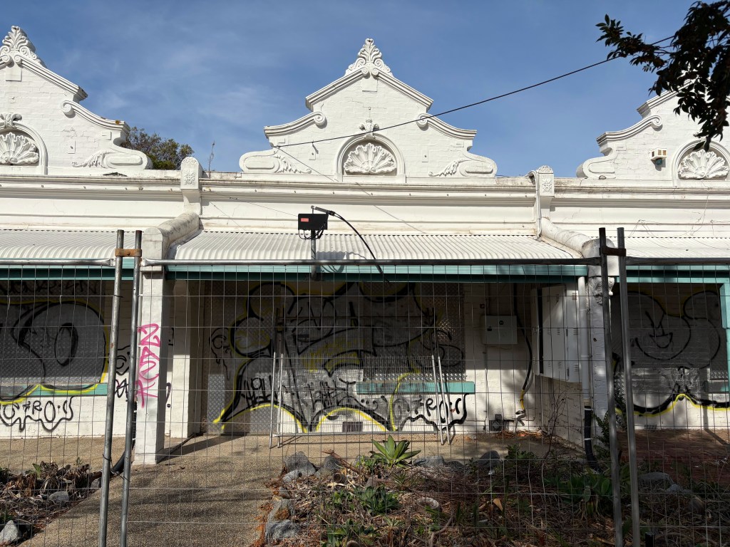 A view of an abandoned building with decorative gable ends, featuring a fence in the foreground and graffiti covering the walls.