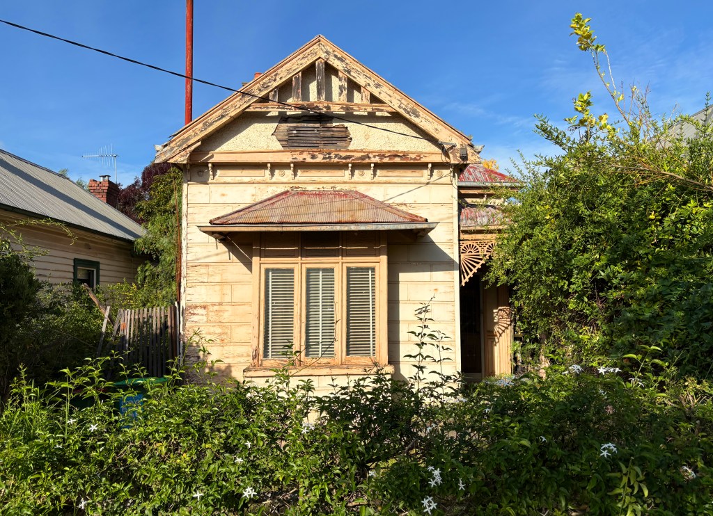 An old, weathered wooden house with a decorative gable and a rusted metal roof, surrounded by overgrown vegetation and flowers.