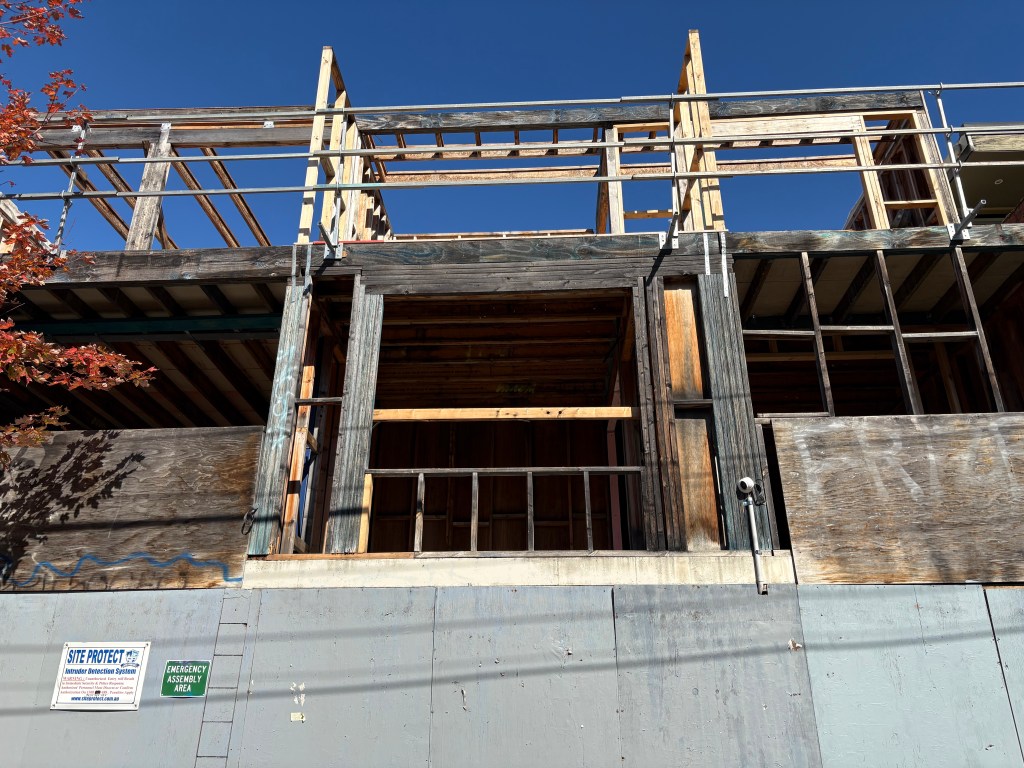 Construction site featuring a partially built wooden structure with scaffolding against a clear blue sky, accompanied by a tree with reddish leaves and safety signs.