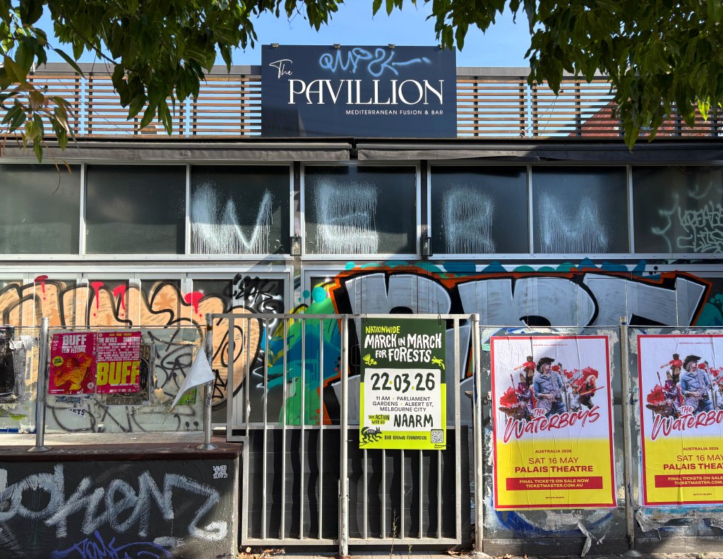 Exterior view of The Pavillion, a Mediterranean fusion restaurant and bar, featuring graffiti on the windows and surrounding walls, with posters advertising events and a green flyer for a forest march.