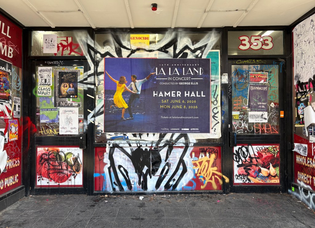 A vibrant street scene showing a shopfront covered in various posters and graffiti, featuring a promotional poster for 'La La Land in Concert' at Hamer Hall, dated June 6 and June 8, 2026.