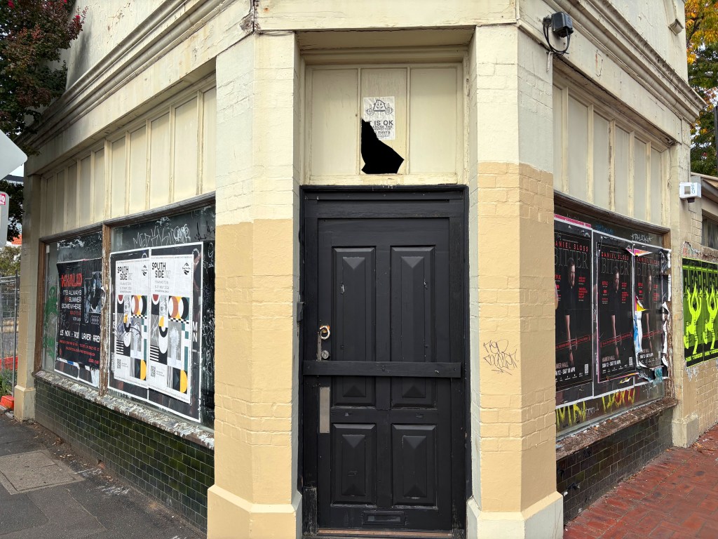 A street corner building with a black door and a broken window. The facade displays several posters for events and shows, with noticeable graffiti on the walls.