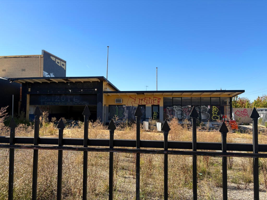 Abandoned building with graffiti and overgrown vegetation, viewed through a black metal fence under a clear blue sky.