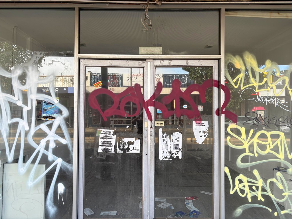 Front entrance of a closed shop covered in various graffiti, including bold tags and stickers on glass doors.
