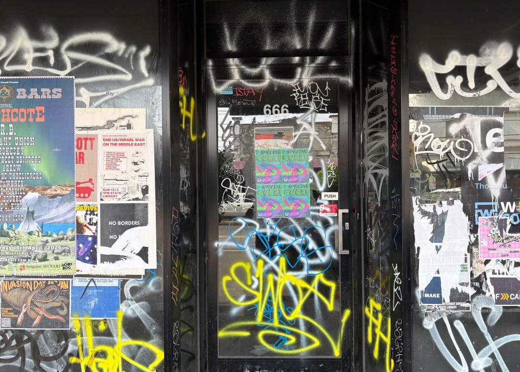 A doorway covered in vibrant graffiti and posters, featuring various event announcements and slogans. The glass door is obscured by layers of spray paint and stickers, adding to the urban aesthetic.