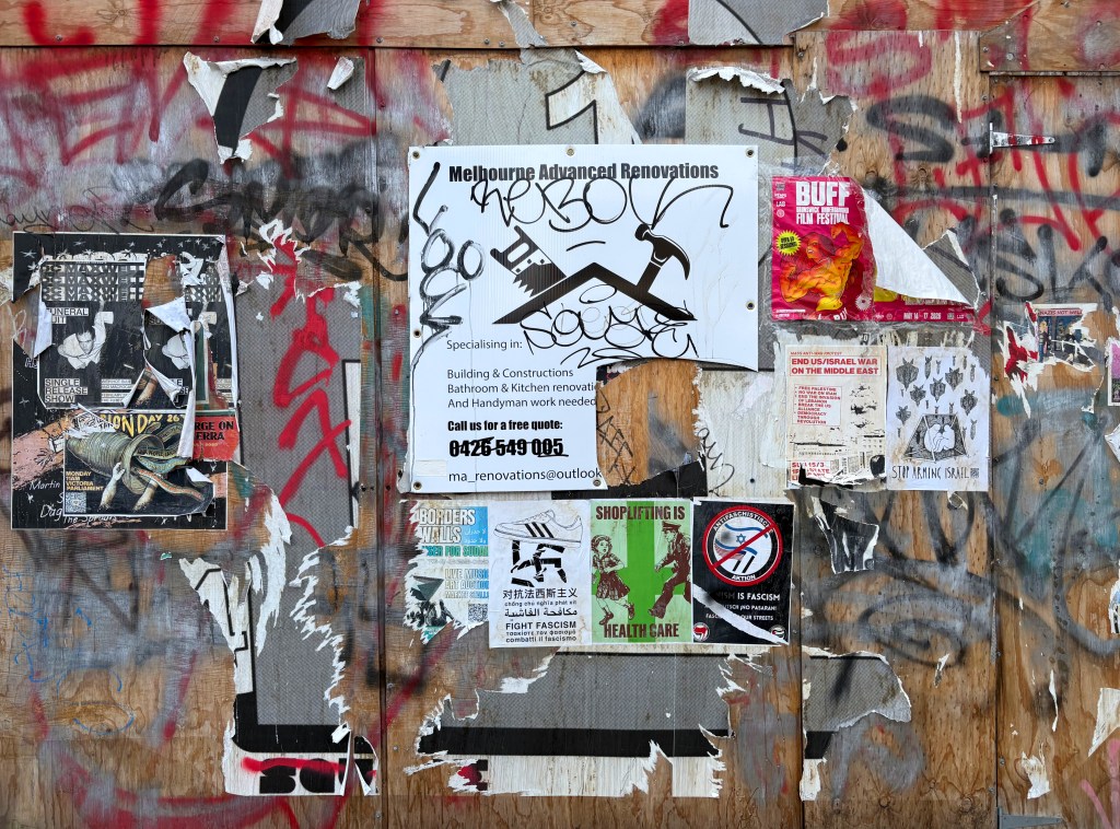 A wooden wall covered with various torn posters, graffiti, and an advertisement for Melbourne Advanced Renovations, featuring contact details and specialisations in construction.