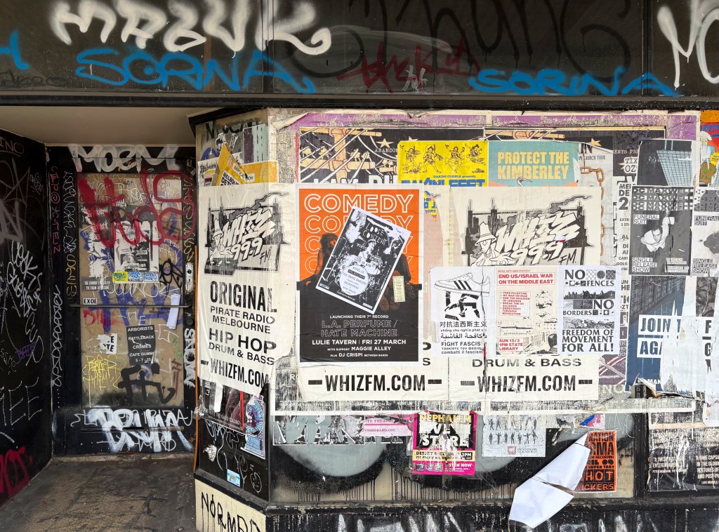 A street corner covered in various posters and graffiti, featuring event advertisements, political messages, and promotional material for local radio and music events.